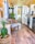 The image depicts a cozy and stylish interior of a tiny home or a mobile home featuring a mix of modern and rustic decor. The walls and ceiling are made of light wood, and the floor has a warm wooden finish. There is a wicker chair with a blue cushion, a wicker basket with a green plant, a small table, and various woven decorations hanging on the walls. The small kitchen area includes white cabinetry, a stove, a refrigerator, a sink, and a wooden countertop. Several plants are placed throughout the space. Towards the back, a bed and more decorations can be seen, with a dog lounging on the bed.