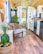 The image depicts a cozy and stylish interior of a tiny home or a mobile home featuring a mix of modern and rustic decor. The walls and ceiling are made of light wood, and the floor has a warm wooden finish. There is a wicker chair with a blue cushion, a wicker basket with a green plant, a small table, and various woven decorations hanging on the walls. The small kitchen area includes white cabinetry, a stove, a refrigerator, a sink, and a wooden countertop. Several plants are placed throughout the space. Towards the back, a bed and more decorations can be seen, with a dog lounging on the bed.
