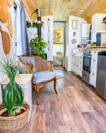 The image depicts a cozy and stylish interior of a tiny home or a mobile home featuring a mix of modern and rustic decor. The walls and ceiling are made of light wood, and the floor has a warm wooden finish. There is a wicker chair with a blue cushion, a wicker basket with a green plant, a small table, and various woven decorations hanging on the walls. The small kitchen area includes white cabinetry, a stove, a refrigerator, a sink, and a wooden countertop. Several plants are placed throughout the space. Towards the back, a bed and more decorations can be seen, with a dog lounging on the bed.