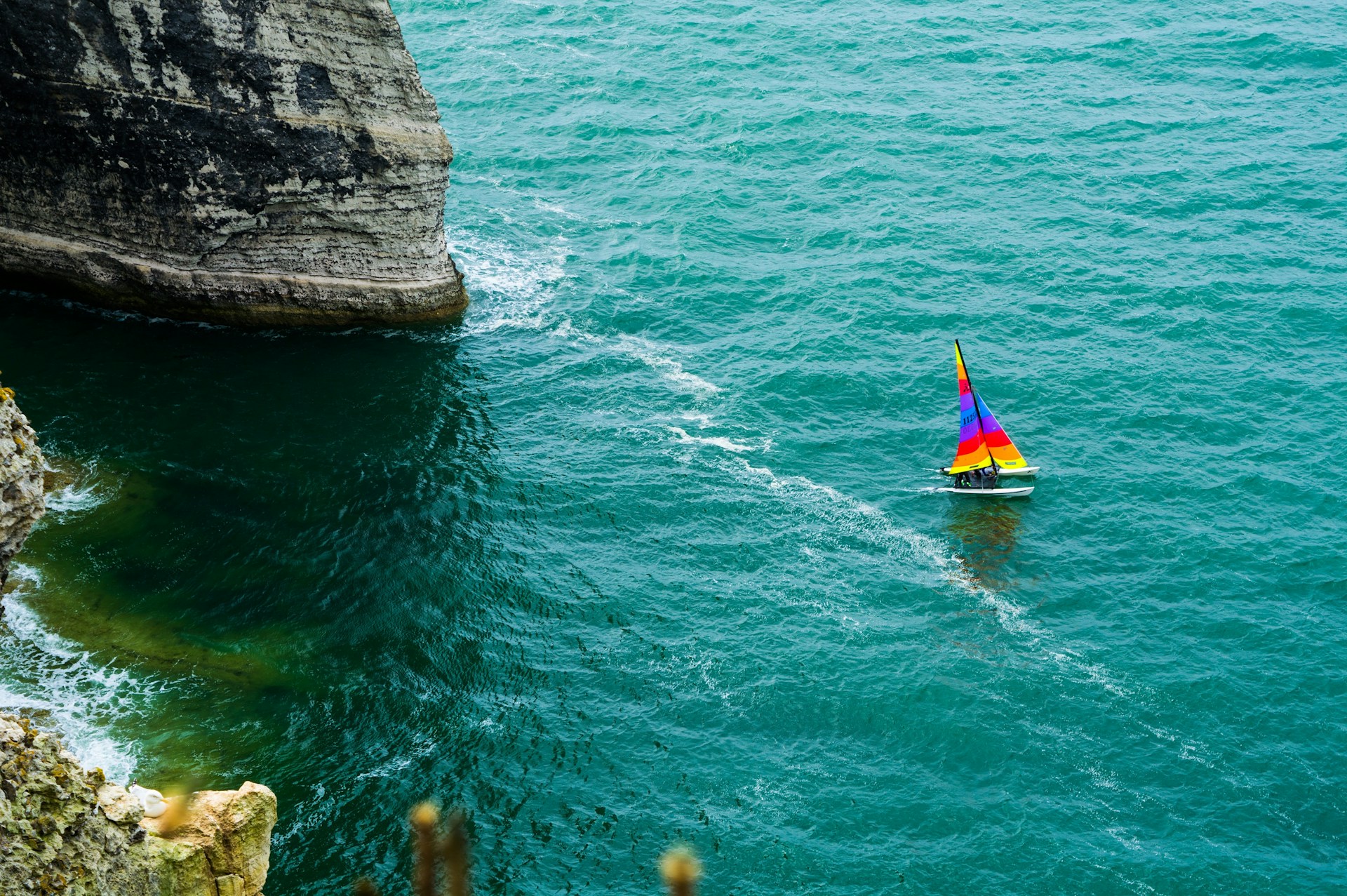 A vibrant sailboat with colorful sails gliding past rocky coastal cliffs on a sunny day.