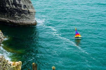 A colorful sailboat navigates the vibrant turquoise waters near steep, rocky cliffs. The sea is slightly choppy, creating a series of white waves behind the sailboat. The sails of the boat display bright hues of red, yellow, and blue, which contrast with the surrounding water and cliffs.