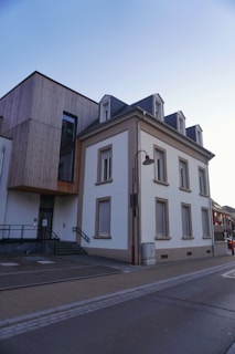 A modern building with a combination of traditional and contemporary architectural elements, featuring a gray mansard roof and wooden paneling on one side. The structure includes large windows and is situated next to a paved street with a sidewalk.