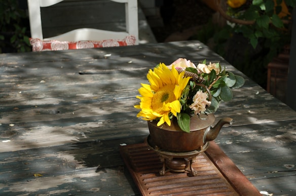 A sunflower and other flowers are arranged in an antique metal teapot on a rustic wooden table. Shadows from surrounding plants create a dappled light effect on the surface. In the background, part of a white chair with a decorative red cushion is visible.