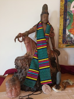A statue of a deity with intricate details sits prominently, adorned with a vibrant, multicolored scarf. The statue is positioned on a decorative base, and in front of it are various objects including a stone head sculpture, a small circular disk with an image, a piece of cord, and a textured object. There is also a colorful tapestry partially visible on the right, adding to the setting&rsquo;s cultural and artistic feel.
