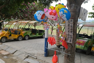A group of colorful character balloons is tied to a tree. Nearby are several small, electric tour vehicles parked on a paved path. Red flags and decorative items hang from the same tree. The setting appears to be a park with greenery in the background.