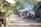 A village street lined with makeshift shops and stalls, with people engaging in daily activities. Some individuals are seen walking, while others appear to be selling or buying goods. The environment is rustic, with natural elements such as trees and greenery framing the scene. Smoke can be seen rising from one of the stalls, suggesting cooking or heating activities.
