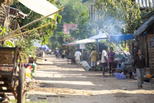 A village street lined with makeshift shops and stalls, with people engaging in daily activities. Some individuals are seen walking, while others appear to be selling or buying goods. The environment is rustic, with natural elements such as trees and greenery framing the scene. Smoke can be seen rising from one of the stalls, suggesting cooking or heating activities.