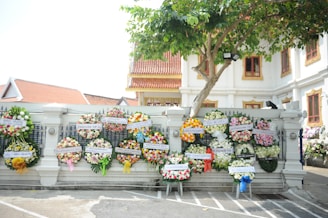 Several colorful floral wreaths are displayed on a white wall in front of a building with traditional architectural elements. A tree with green leaves partially shades the area. The setting appears to be part of a cultural or ceremonial site.