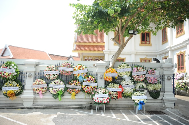 Several colorful floral wreaths are displayed on a white wall in front of a building with traditional architectural elements. A tree with green leaves partially shades the area. The setting appears to be part of a cultural or ceremonial site.