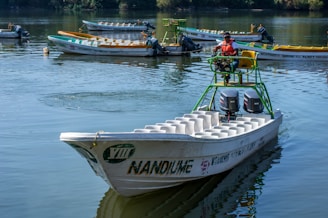 A man wearing a life jacket is operating a motorboat on a tranquil body of water. The boat is equipped with two outboard engines and has an open seating layout. Several similar boats are visible in the background, moored and reflecting the surrounding trees on the water's surface.