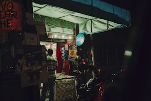 A small storefront illuminated by artificial light during nighttime. A person in a striped shirt is seen entering, with various products displayed inside. The entrance is decorated with advertising posters, and a scooter is parked nearby.