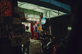 A small storefront illuminated by artificial light during nighttime. A person in a striped shirt is seen entering, with various products displayed inside. The entrance is decorated with advertising posters, and a scooter is parked nearby.