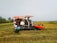 A rice field where a harvester is actively working. Several sacks of harvested grain are visible piled onto the machine. Workers are engaged in the process, with some standing nearby, likely overseeing the operation in the background. The scene is set in a rural landscape with open skies and distant trees lining the horizon.