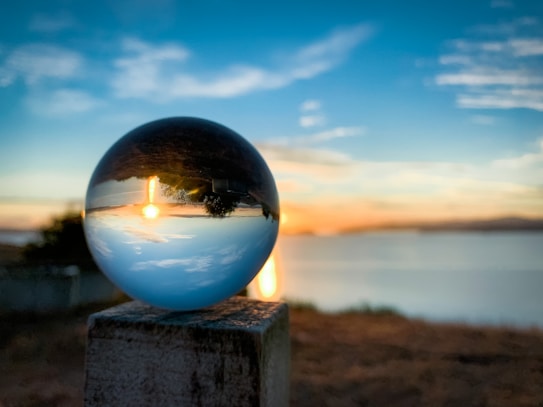 A crystal ball placed on a wooden post reflects a vivid sunset over a calm body of water. The sky is dramatic with a mix of orange and blue hues, and the reflection in the sphere creates an inverted effect, with the bright sun near the horizon and clouds scattered across the sky.