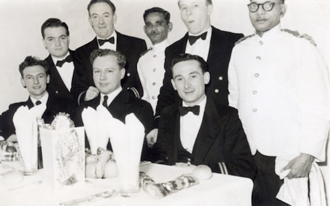 A group of men dressed in formal attire, seated at a dining table with white tablecloths and napkins. Some are wearing tuxedos or dinner jackets while others appear to be in uniform. The setting suggests a formal or celebratory event, with visible dining ware and decorations on the table.