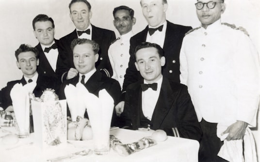 A group of men dressed in formal attire, seated at a dining table with white tablecloths and napkins. Some are wearing tuxedos or dinner jackets while others appear to be in uniform. The setting suggests a formal or celebratory event, with visible dining ware and decorations on the table.