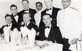 A group of men dressed in formal attire, seated at a dining table with white tablecloths and napkins. Some are wearing tuxedos or dinner jackets while others appear to be in uniform. The setting suggests a formal or celebratory event, with visible dining ware and decorations on the table.
