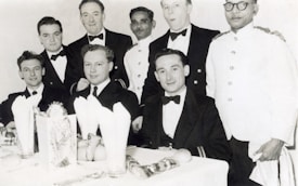 A group of men dressed in formal attire, seated at a dining table with white tablecloths and napkins. Some are wearing tuxedos or dinner jackets while others appear to be in uniform. The setting suggests a formal or celebratory event, with visible dining ware and decorations on the table.