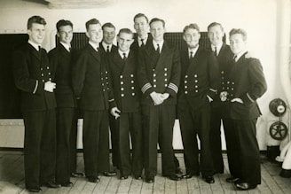 A group of ten men in uniform stand in close proximity on a wooden deck. They are wearing formal naval attire with double-breasted jackets, some adorned with stripes on their sleeves. Their expressions are cheerful, suggesting camaraderie.