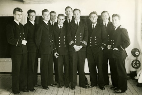 A group of ten men in uniform stand in close proximity on a wooden deck. They are wearing formal naval attire with double-breasted jackets, some adorned with stripes on their sleeves. Their expressions are cheerful, suggesting camaraderie.