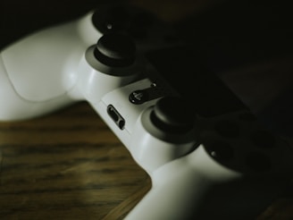 A white gaming controller rests on a wooden surface. The lighting is low, casting shadows across the controller, highlighting its buttons and joysticks.
