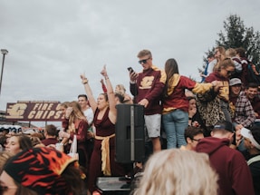 A lively crowd of young people wearing maroon and gold clothing gather at an outdoor event. Many are cheering, smiling, and holding drinks. A large sign in the background reads 'FIRE CHIPS'. The sky is overcast and there are trees and a lamppost visible.