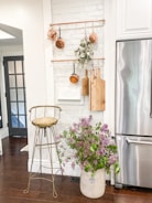 A cozy kitchen corner with a white textured brick wall displaying copper pots and wooden cutting boards hanging from brass rods. Below, a rustic metal stool stands next to a large ceramic pot filled with lilac flowers. A stainless steel refrigerator is visible to the right, and a wooden floor enhances the warm ambiance.