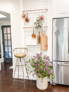 A cozy kitchen corner with a white textured brick wall displaying copper pots and wooden cutting boards hanging from brass rods. Below, a rustic metal stool stands next to a large ceramic pot filled with lilac flowers. A stainless steel refrigerator is visible to the right, and a wooden floor enhances the warm ambiance.