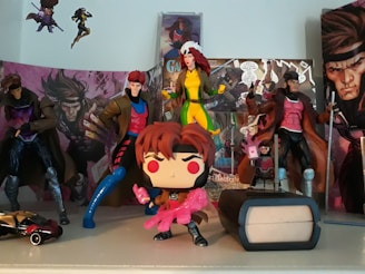 A collection of superhero action figures and collectibles displayed on a shelf. The figures are dressed in various colorful costumes, and the backdrop includes comic book covers featuring dynamic art. A small toy car and a mini figurine of a character in a trench coat decorate the scene.