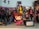 A collection of superhero action figures and collectibles displayed on a shelf. The figures are dressed in various colorful costumes, and the backdrop includes comic book covers featuring dynamic art. A small toy car and a mini figurine of a character in a trench coat decorate the scene.