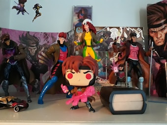A collection of superhero action figures and collectibles displayed on a shelf. The figures are dressed in various colorful costumes, and the backdrop includes comic book covers featuring dynamic art. A small toy car and a mini figurine of a character in a trench coat decorate the scene.