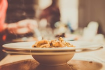 A plate of pasta with mushrooms is placed on a wooden table. The focus is on the food, which seems to be freshly cooked, while the background is blurred, suggesting a kitchen setting.