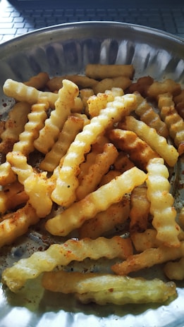 A close-up view of crinkle-cut fries seasoned and served in a round metallic plate. The fries are golden-brown, indicating they've been cooked to a crispy texture. Light from the sun highlights parts of the fries and plate, adding warmth to the scene.