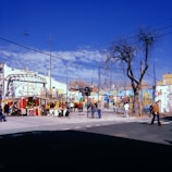 A street scene featuring a colorful market against a backdrop of buildings with vibrant murals. The market stalls are filled with various items, and several people are browsing or walking around. The sky is clear and blue, and tree branches are visible, some leafless suggesting a cool season.