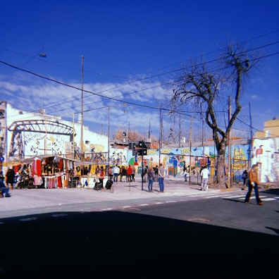 A street scene featuring a colorful market against a backdrop of buildings with vibrant murals. The market stalls are filled with various items, and several people are browsing or walking around. The sky is clear and blue, and tree branches are visible, some leafless suggesting a cool season.