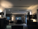A dimly lit, modern living room with dark furniture and decor. The space is framed by brick walls with two narrow windows featuring horizontal blinds. Several lamps provide warm accent lighting, and a rug anchors a seating area with chairs. The overall style is minimalist and contemporary.