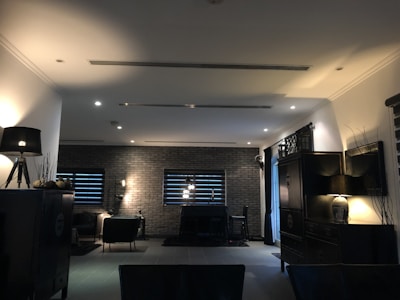 A dimly lit, modern living room with dark furniture and decor. The space is framed by brick walls with two narrow windows featuring horizontal blinds. Several lamps provide warm accent lighting, and a rug anchors a seating area with chairs. The overall style is minimalist and contemporary.