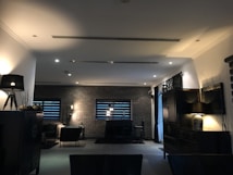 A dimly lit, modern living room with dark furniture and decor. The space is framed by brick walls with two narrow windows featuring horizontal blinds. Several lamps provide warm accent lighting, and a rug anchors a seating area with chairs. The overall style is minimalist and contemporary.