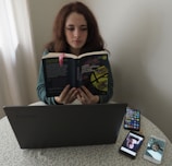 A person sitting at a table reading a book titled 'Cent'anni di solitudine' by Gabriel Garcia Marquez. In front of them is a laptop on the table, alongside two smartphones displaying images. The setting appears to be indoors, with a light-colored curtain in the background.