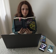 A person sitting at a table reading a book titled 'Cent'anni di solitudine' by Gabriel Garcia Marquez. In front of them is a laptop on the table, alongside two smartphones displaying images. The setting appears to be indoors, with a light-colored curtain in the background.