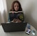 A person sitting at a table reading a book titled 'Cent'anni di solitudine' by Gabriel Garcia Marquez. In front of them is a laptop on the table, alongside two smartphones displaying images. The setting appears to be indoors, with a light-colored curtain in the background.