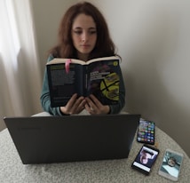 A person sitting at a table reading a book titled 'Cent'anni di solitudine' by Gabriel Garcia Marquez. In front of them is a laptop on the table, alongside two smartphones displaying images. The setting appears to be indoors, with a light-colored curtain in the background.