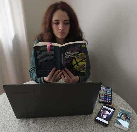 A person sitting at a table reading a book titled 'Cent'anni di solitudine' by Gabriel Garcia Marquez. In front of them is a laptop on the table, alongside two smartphones displaying images. The setting appears to be indoors, with a light-colored curtain in the background.