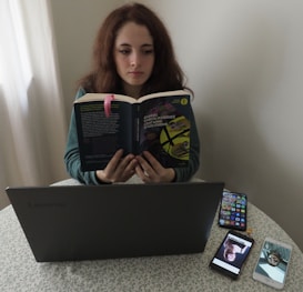 A person sitting at a table reading a book titled 'Cent'anni di solitudine' by Gabriel Garcia Marquez. In front of them is a laptop on the table, alongside two smartphones displaying images. The setting appears to be indoors, with a light-colored curtain in the background.