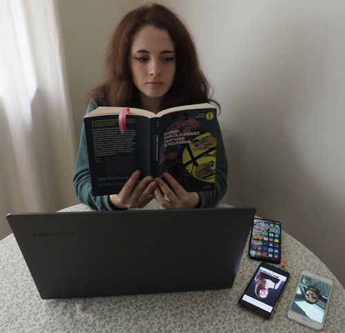 A person sitting at a table reading a book titled 'Cent'anni di solitudine' by Gabriel Garcia Marquez. In front of them is a laptop on the table, alongside two smartphones displaying images. The setting appears to be indoors, with a light-colored curtain in the background.