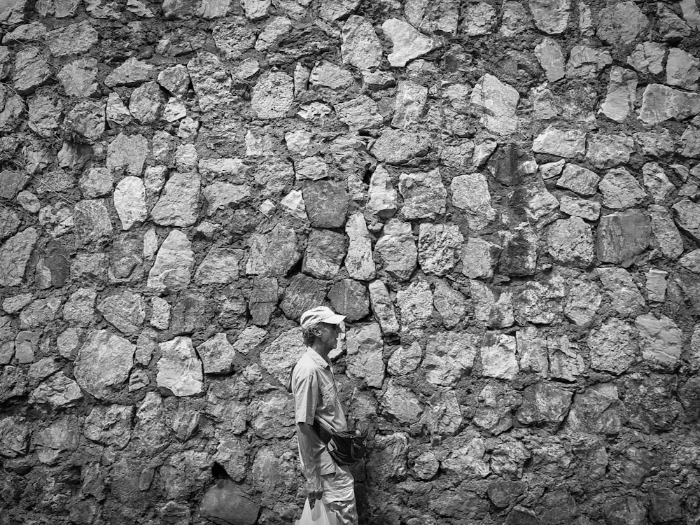 grayscale photo of man in long sleeve shirt and pants standing on brick wall