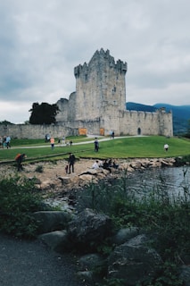 A historic stone castle stands prominently against a backdrop of cloudy skies and distant mountains. People are walking and sitting by the water's edge, surrounded by lush greenery and stones. The castle appears to have tall, narrow windows and battlements, indicative of a medieval architectural style.