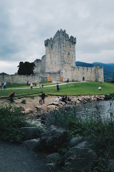A historic stone castle stands prominently against a backdrop of cloudy skies and distant mountains. People are walking and sitting by the water's edge, surrounded by lush greenery and stones. The castle appears to have tall, narrow windows and battlements, indicative of a medieval architectural style.