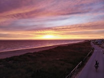 A serene beach scene at sunset with vibrant colors in the sky and gentle waves lapping the shore. A solitary rider on horseback can be seen on the road alongside the dunes, heading away from the ocean. The coastline extends into the distance, lined with a few small buildings or houses.