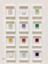 A grid of twelve small framed squares, each containing a single colored square. The frames are uniformly white, arranged in three columns and four rows on a plain wall. Each square is a distinct color, ranging from pastel and neutral tones to bold and vibrant hues.